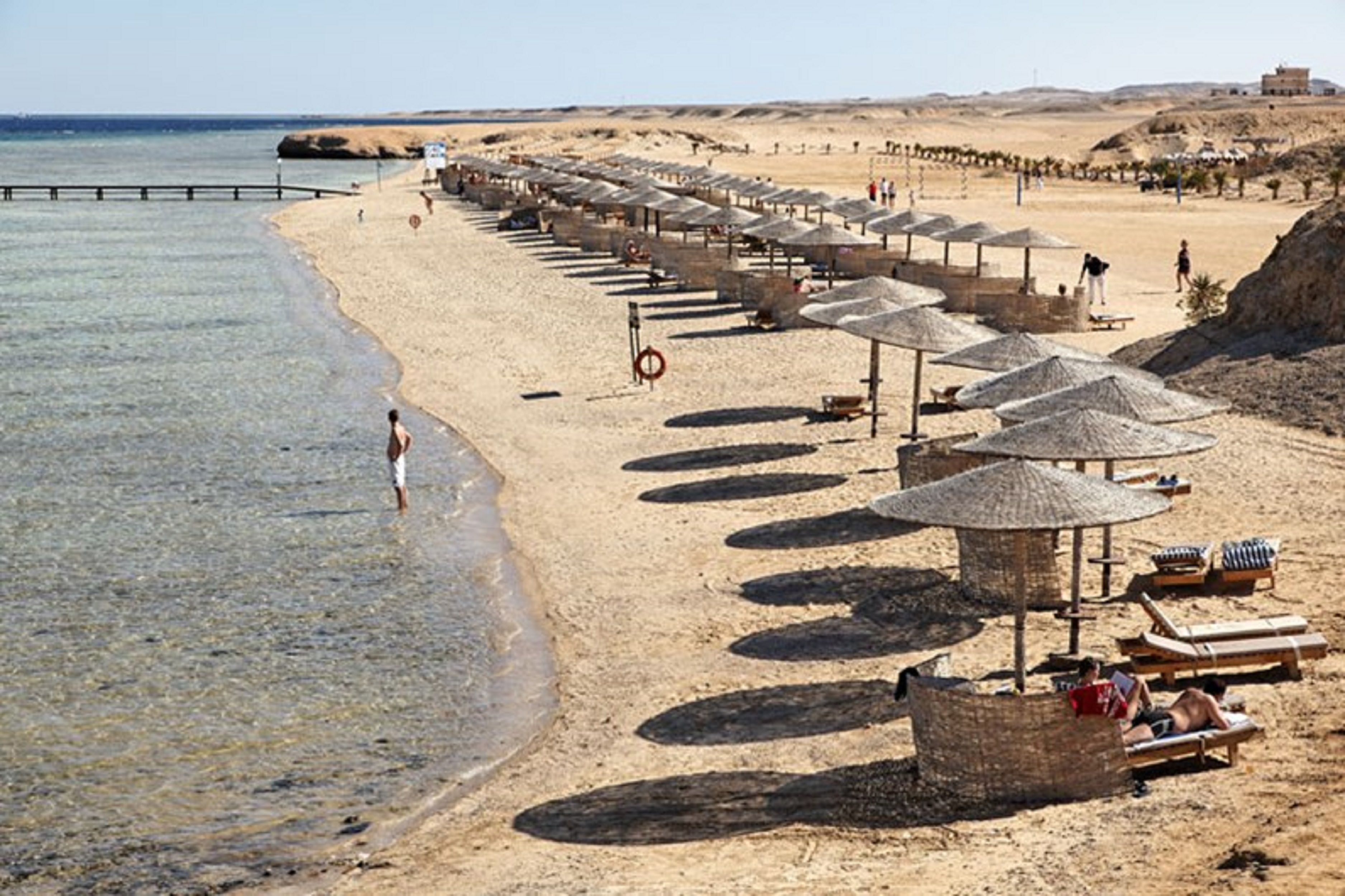 A beach with rows of sun umbrellas and lounge chairs, a clear view of the sea, and a distant building on the horizon.