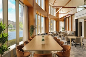 A spacious conference room with a long table and multiple chairs, large windows with curtains, and indoor plants.