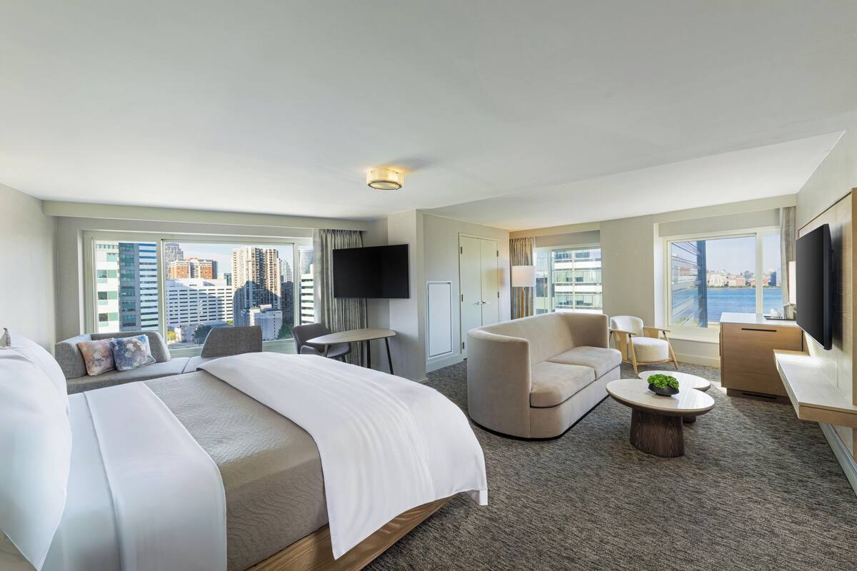 A hotel room with a large bed, a sofa, a coffee table, and a TV. There are windows with a city view.