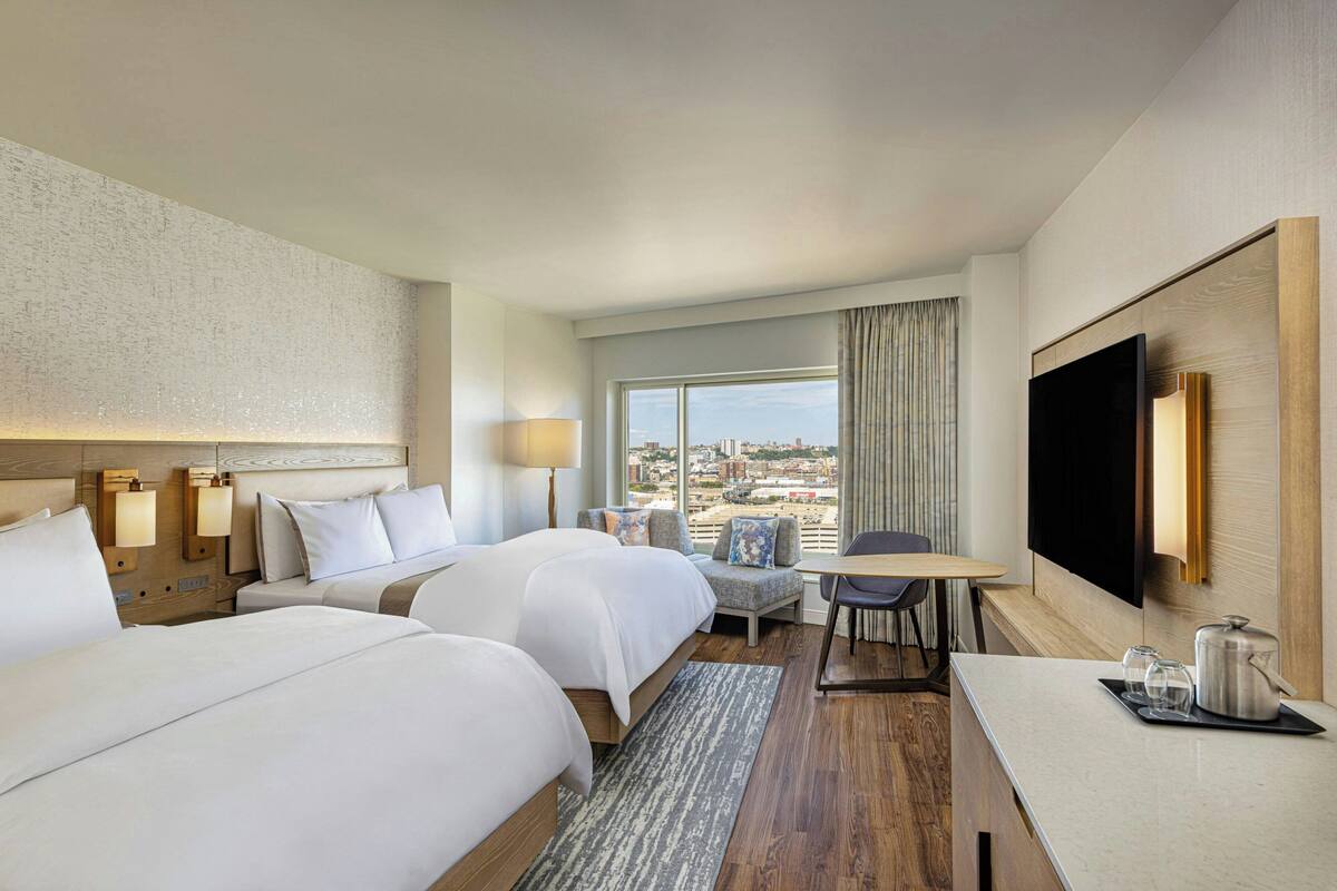 A hotel room with two beds, a desk, a TV, and a view of the city.