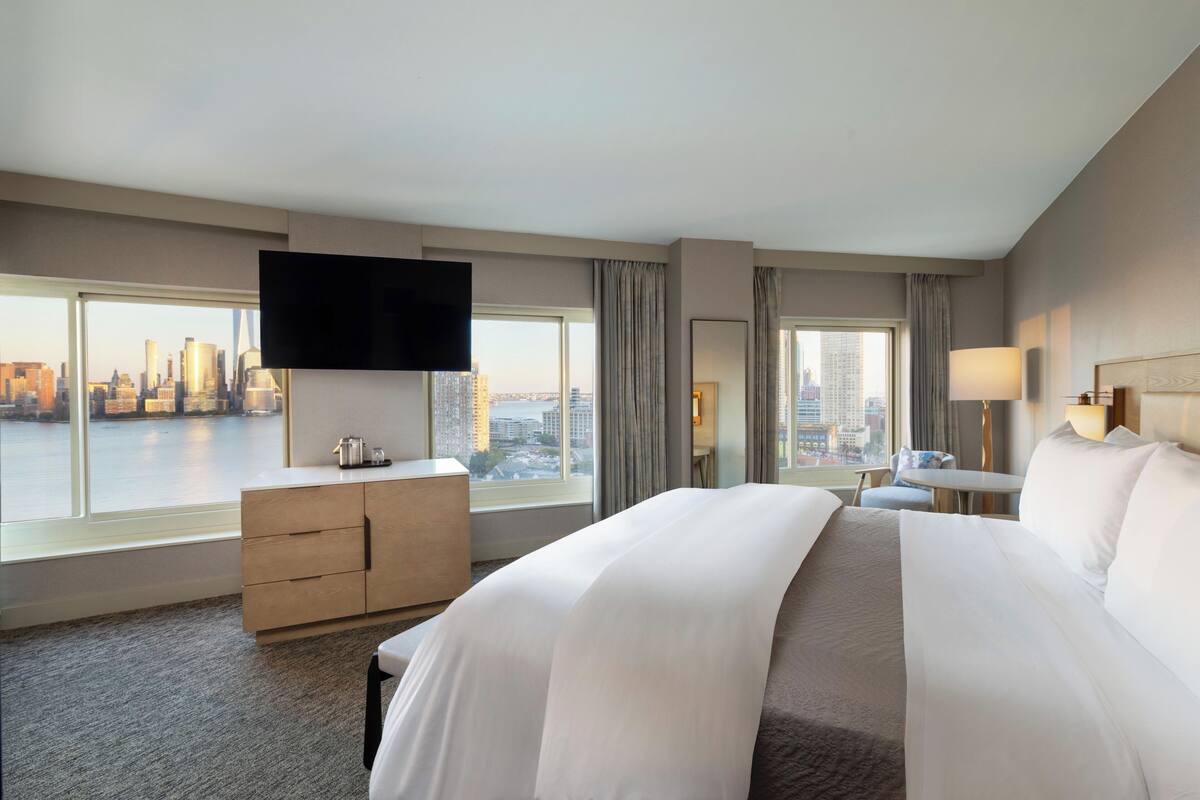 A hotel room with a large bed, a TV mounted on the wall, a desk with a chair, and a view of the cityscape through large windows.