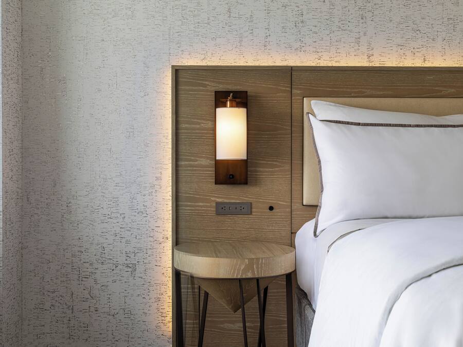 Un lit bien fait, avec du linge de lit blanc, une tête de lit en bois et une petite table de chevet sur laquelle se trouve une lampe.