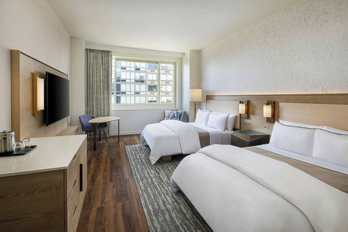 A hotel room with two beds, a desk, a TV, a window with curtains, and a view of buildings.