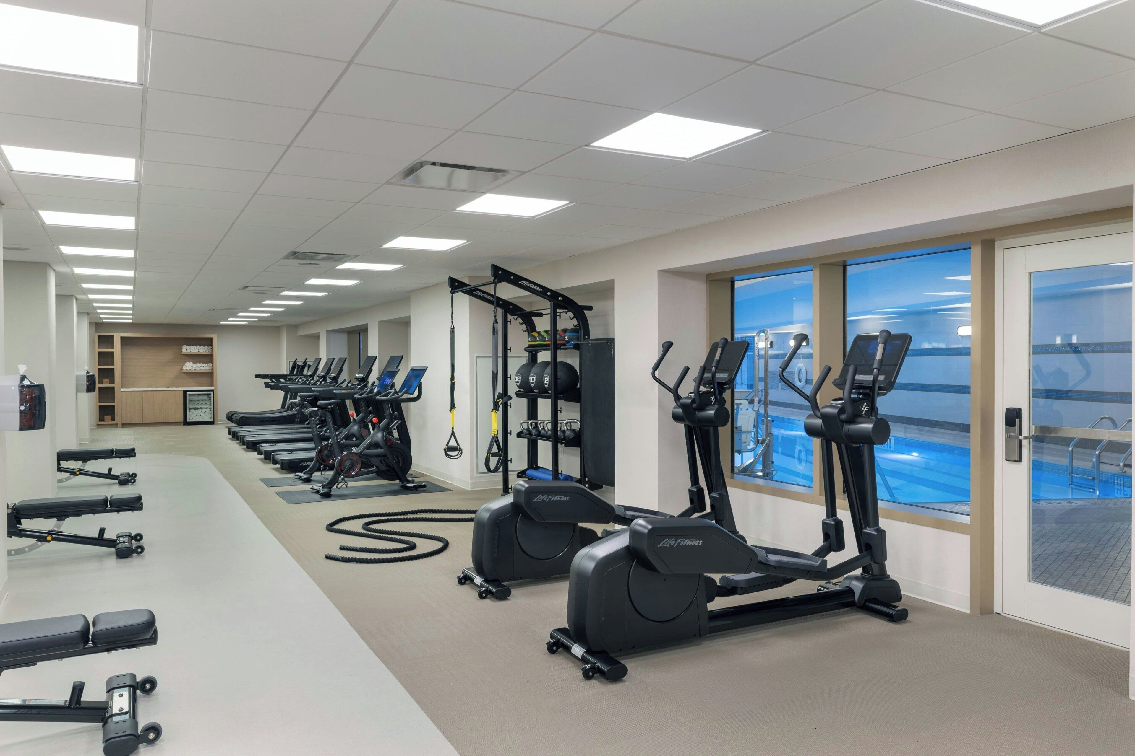 A gym with various cardio machines, weightlifting equipment, and a large window with a view of a pool.