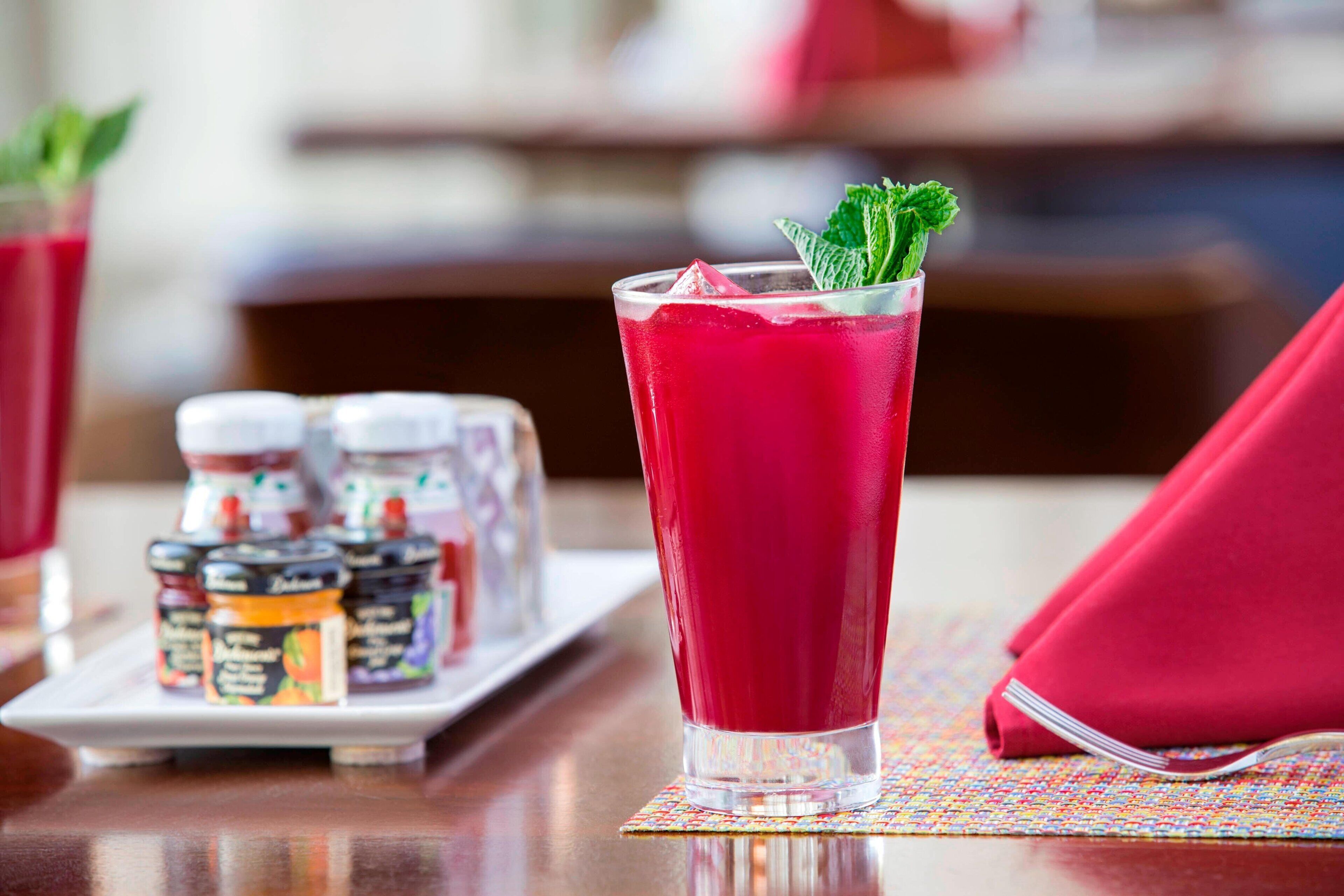 A glass of red beverage with mint garnish on a table, accompanied by a tray of condiments and a folded napkin.