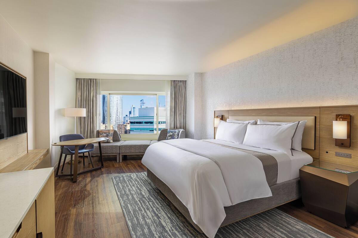 A hotel room with a large bed, a desk, a chair, a small table, and a view of the city.