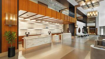 A modern hotel lobby with a reception desk, seating area, and a staircase leading to upper floors.