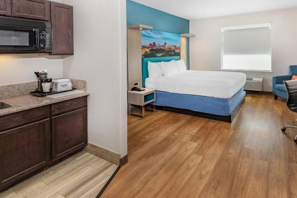 A compact hotel room with a kitchenette, a bed, a desk, and a window.