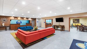 A hotel lobby with a reception desk, a blue sofa, a red sofa, a dining area with tables and chairs, and a TV mounted on the wall. - Days Inn & Suites by Wyndham San Antonio near Frost Bank Center (San Antonio)