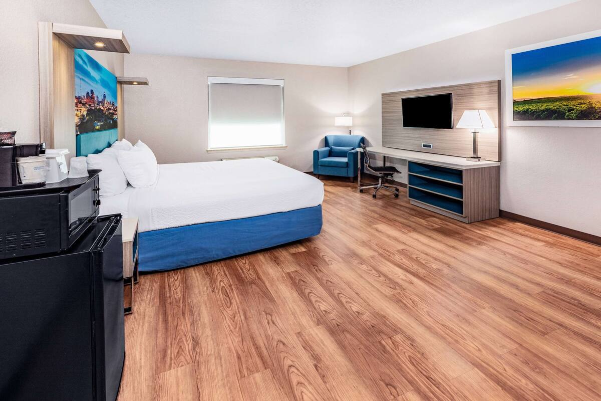 A hotel room with a bed, a desk with a TV, a chair, and a painting on the wall.