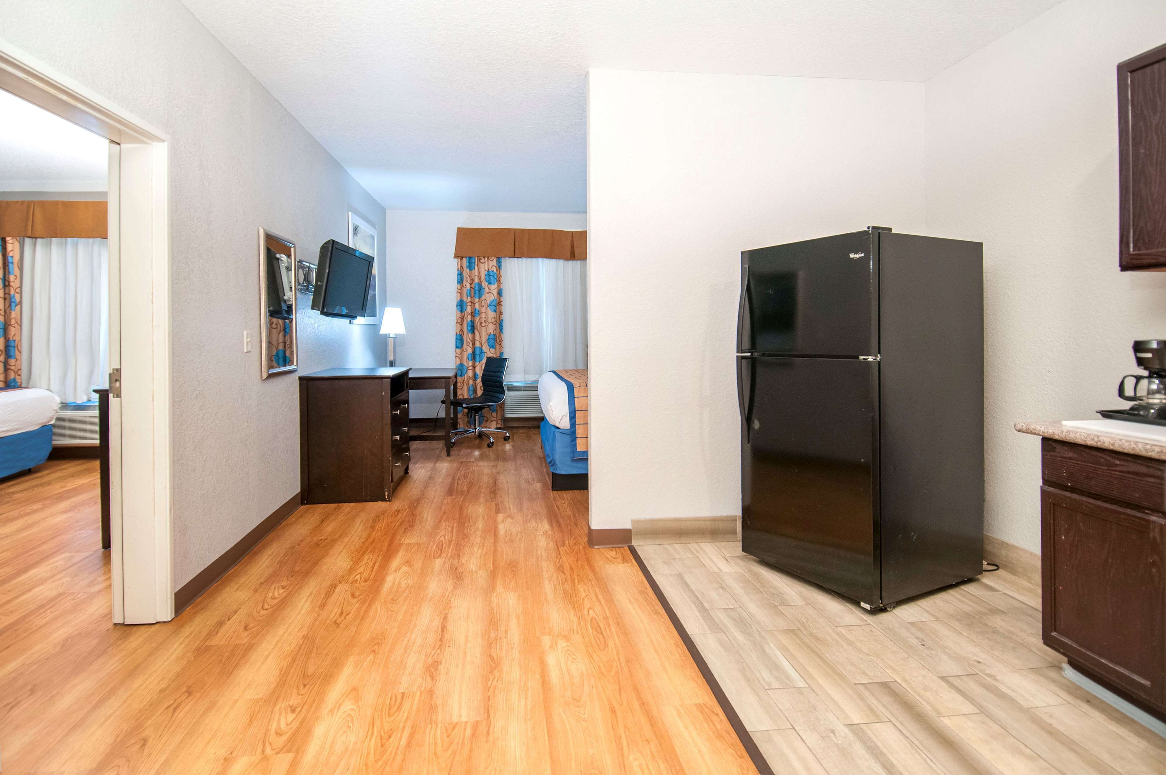 A hotel room with a wooden floor, a black refrigerator, a desk with a chair, a television, and a bed with a blue cover.