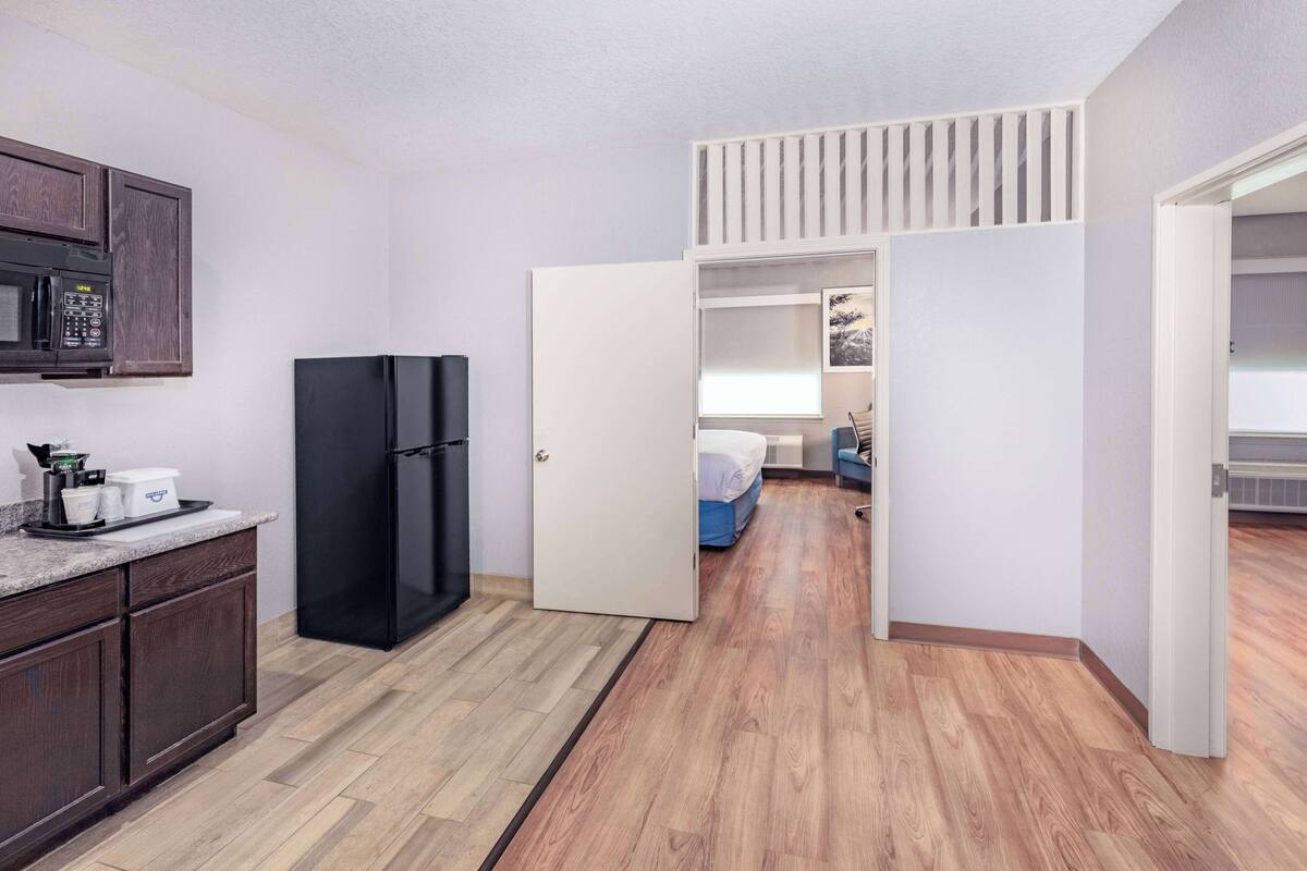 A compact kitchen with a microwave, refrigerator, and a small countertop with kitchenware. A bedroom with a bed and a window is visible through an open door.