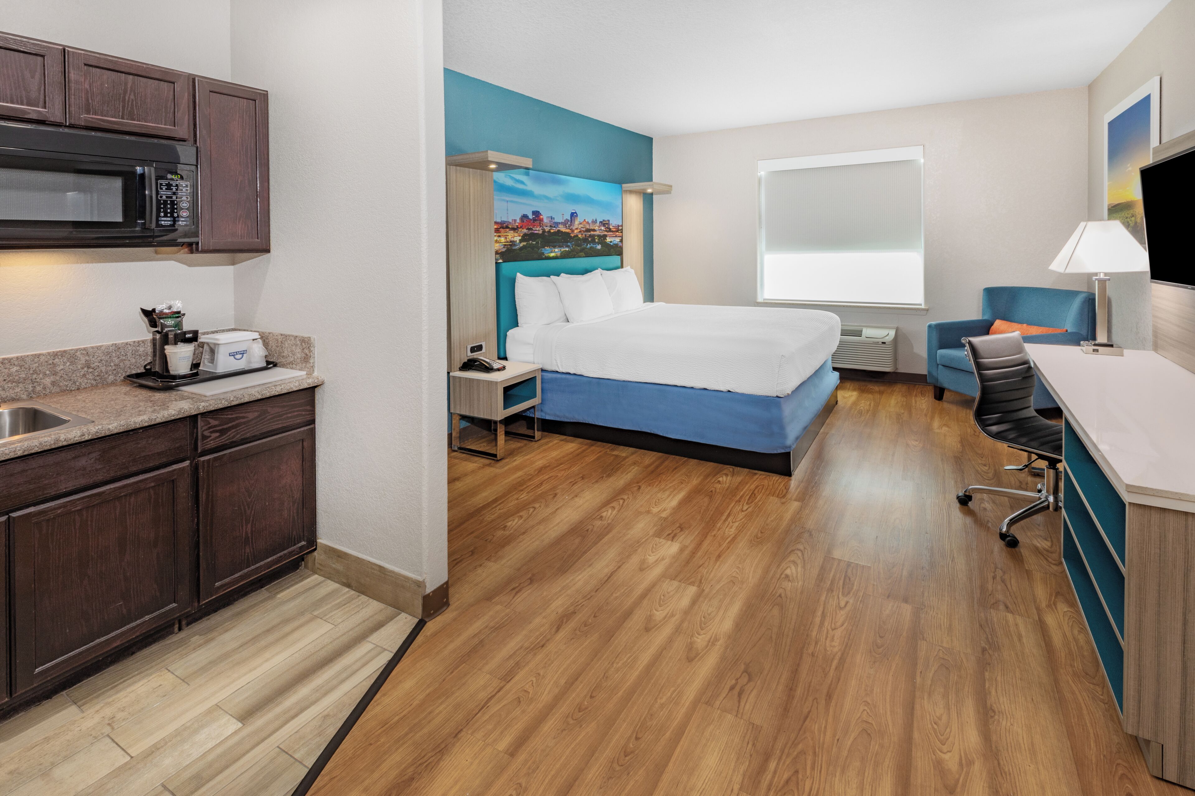 A hotel room with a bed, a desk, a chair, a microwave, a coffee maker, and a painting on the wall.