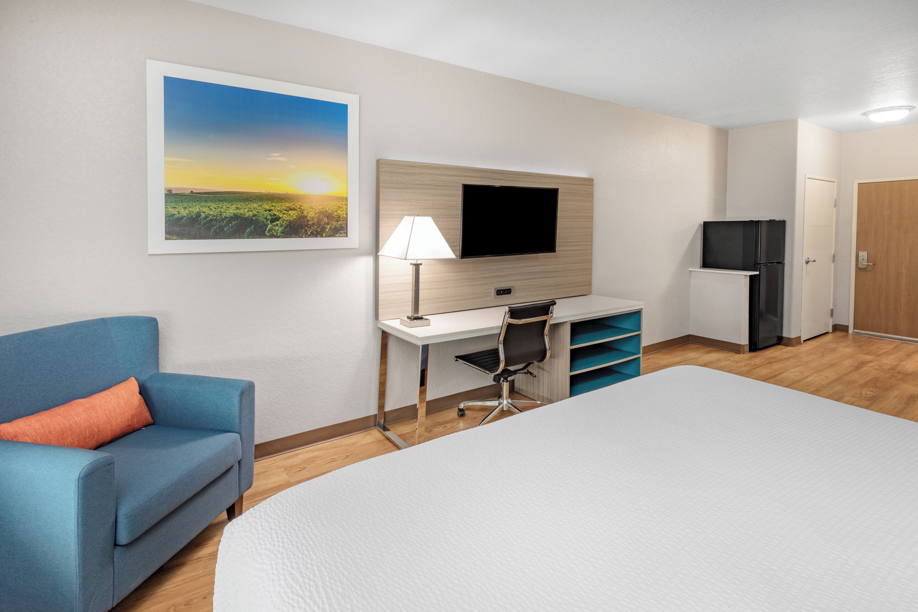 A hotel room with a bed, a desk with a TV, a sofa, a refrigerator, and a door.