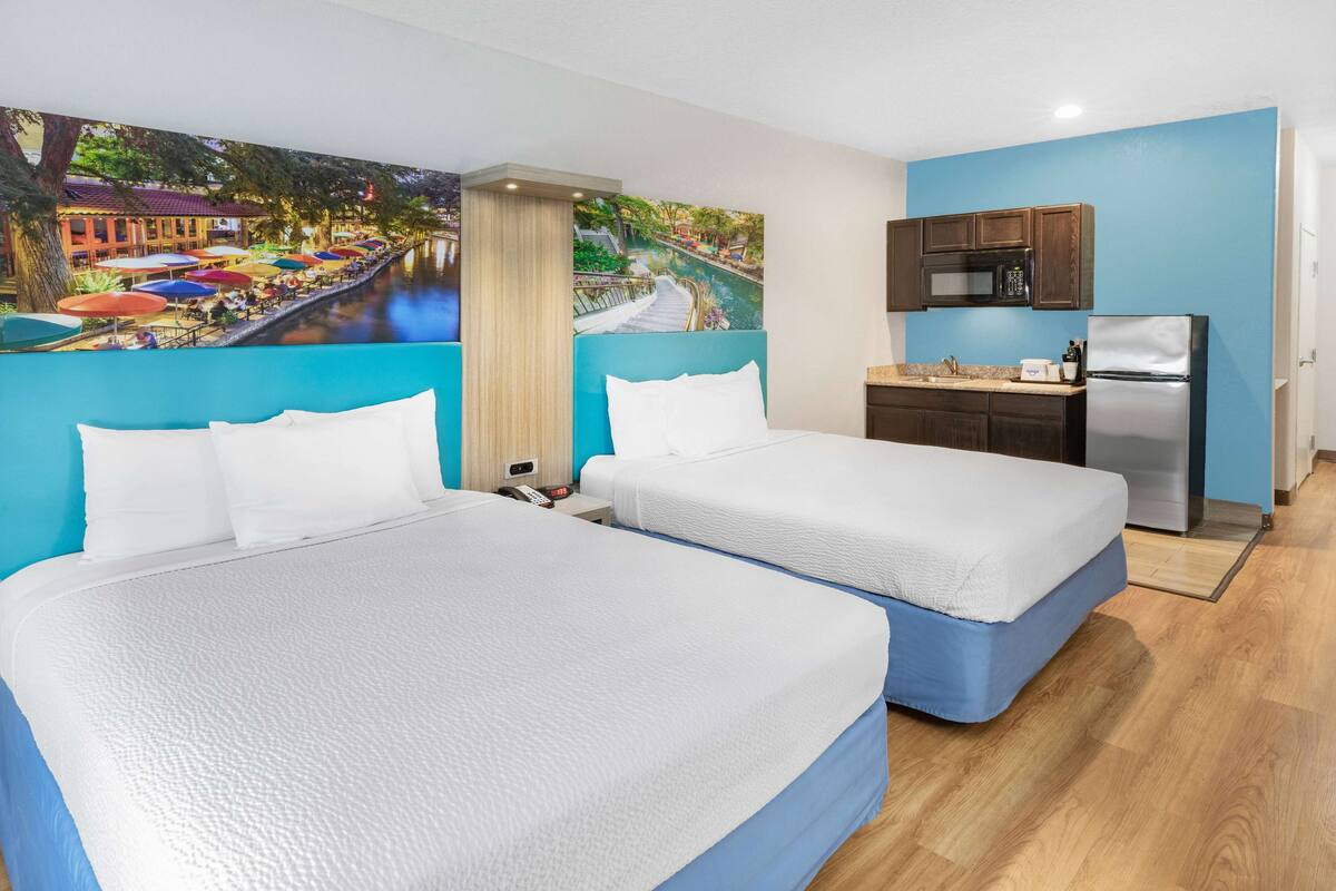 A hotel room with two beds, a kitchenette, and a mural of a riverside scene.