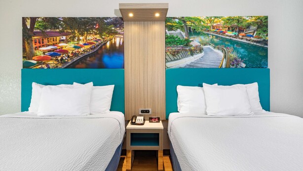 A hotel room with two beds, a nightstand with a phone and alarm clock, and a large mural of a riverside scene. - Days Inn & Suites by Wyndham San Antonio near Frost Bank Center (San Antonio)