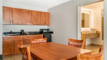 A compact kitchen with wooden cabinets, a microwave, a coffee maker, and a refrigerator. There is a dining table with chairs and a patterned cushion.