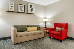 A hotel room with a beige sofa, a red armchair, a small table with a lamp, and three framed artworks on the wall. - Country Inn & Suites by Radisson, Rossford - Toledo South (Perrysburg)