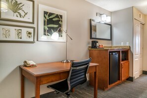 A hotel room with a wooden desk, a black office chair, a desk lamp, a minibar, a refrigerator, a mirror, and framed botanical artwork on the wall. - Country Inn & Suites by Radisson, Rossford - Toledo South (Perrysburg)