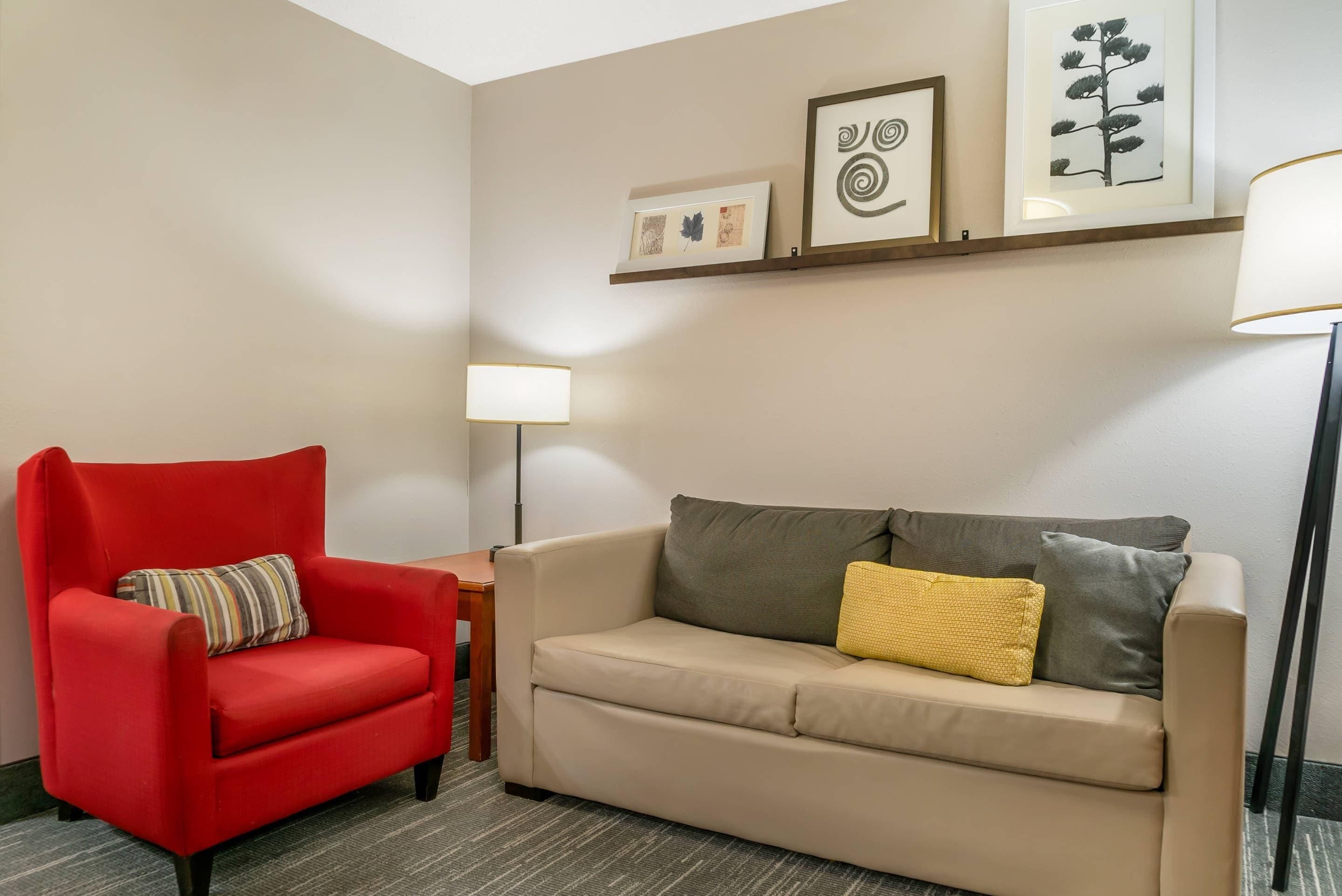 A room with a red armchair, a beige sofa, a small table, and a floor lamp. There are three framed artworks on the wall.