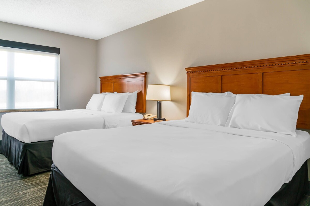 Habitación de hotel con dos camas, una mesita de noche con lámpara y una ventana con persianas.