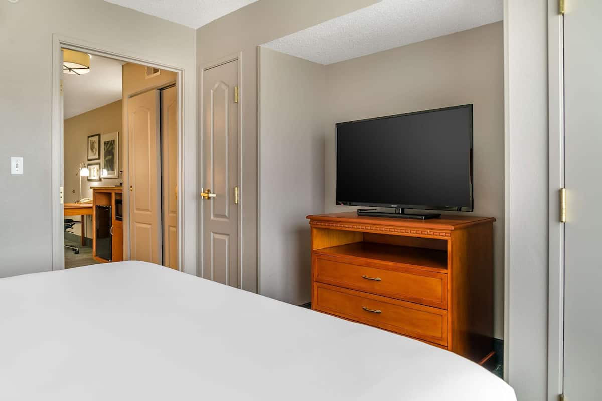 A bedroom with a bed, a wooden dresser, a television, and a door leading to a bathroom.