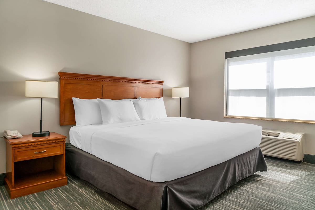 A hotel room with a large bed, two bedside lamps, a nightstand with a phone, and an air conditioning unit.