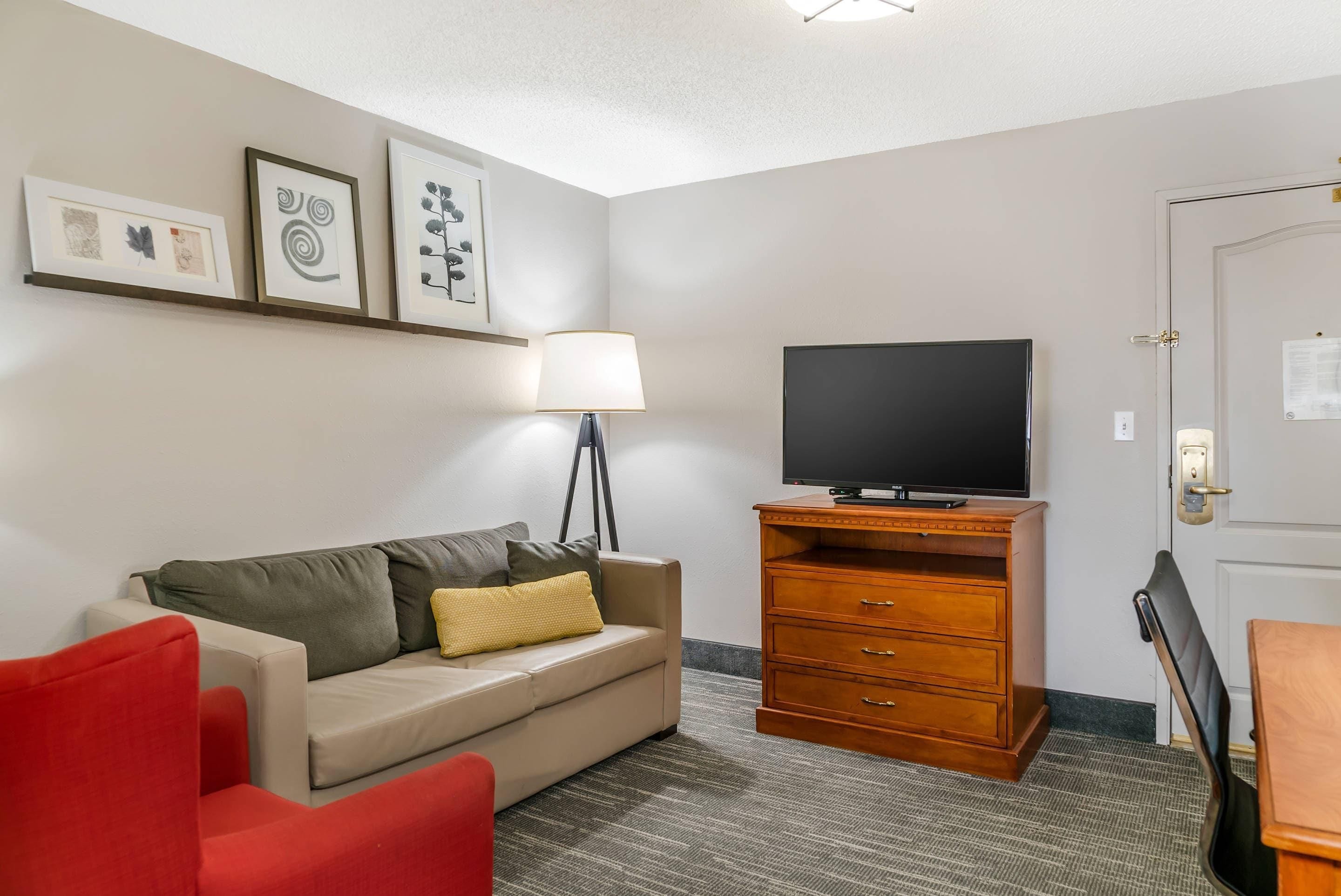 Quarto de hotel com sofá, uma cadeira vermelha, TV sobre um suporte de madeira, abajur e quadros nas paredes.