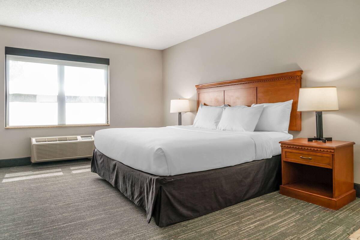 A hotel room with a large bed, two bedside lamps, a nightstand, and a window with blinds.