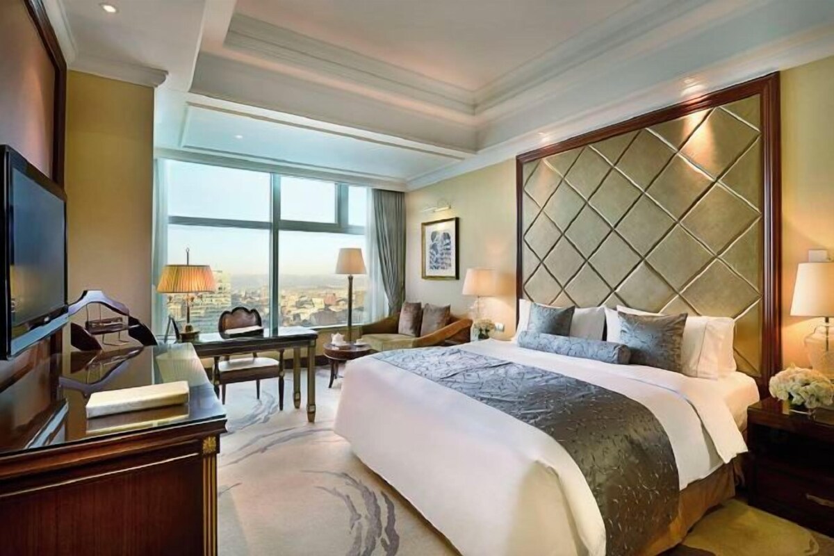 A hotel room with a large bed, a television, a desk with a chair, and a view of the city through large windows.