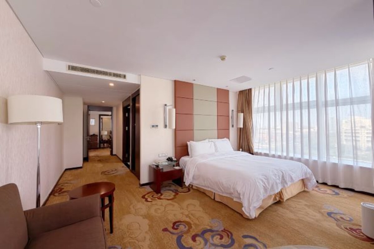 A hotel room with a bed, a desk, a chair, a lamp, and a large window.