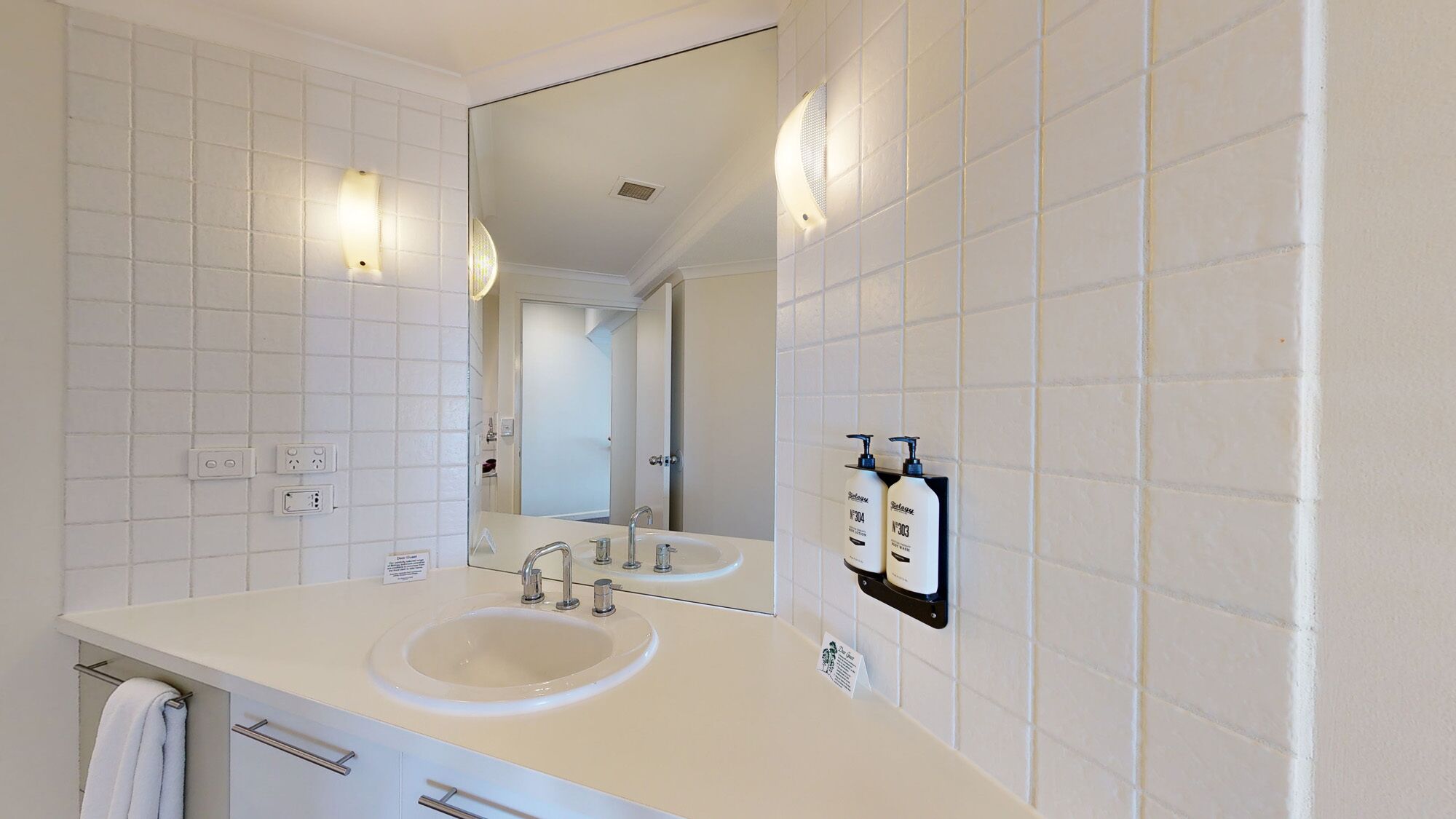 Une salle de bain moderne avec des murs recouverts de carreaux blancs, un grand miroir, un lavabo double et deux bouteilles de shampoing.
