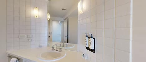 Une salle de bain moderne avec des murs recouverts de carreaux blancs, un grand miroir, un lavabo double et deux bouteilles de shampoing.