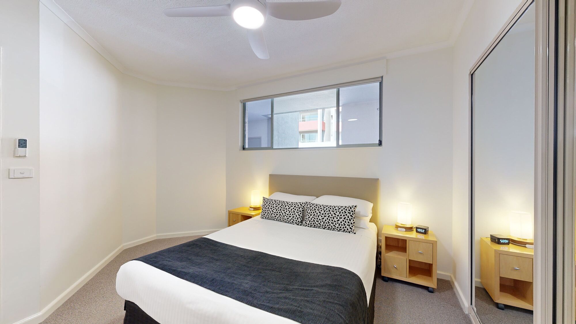 A hotel room with a bed, bedside tables, a ceiling fan, and a window with a view of buildings.
