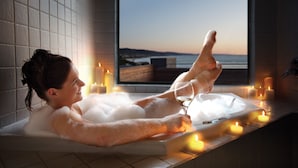 Uma pessoa relaxando em uma banheira com velas e uma taça de vinho, com vista para o mar através de uma janela.
