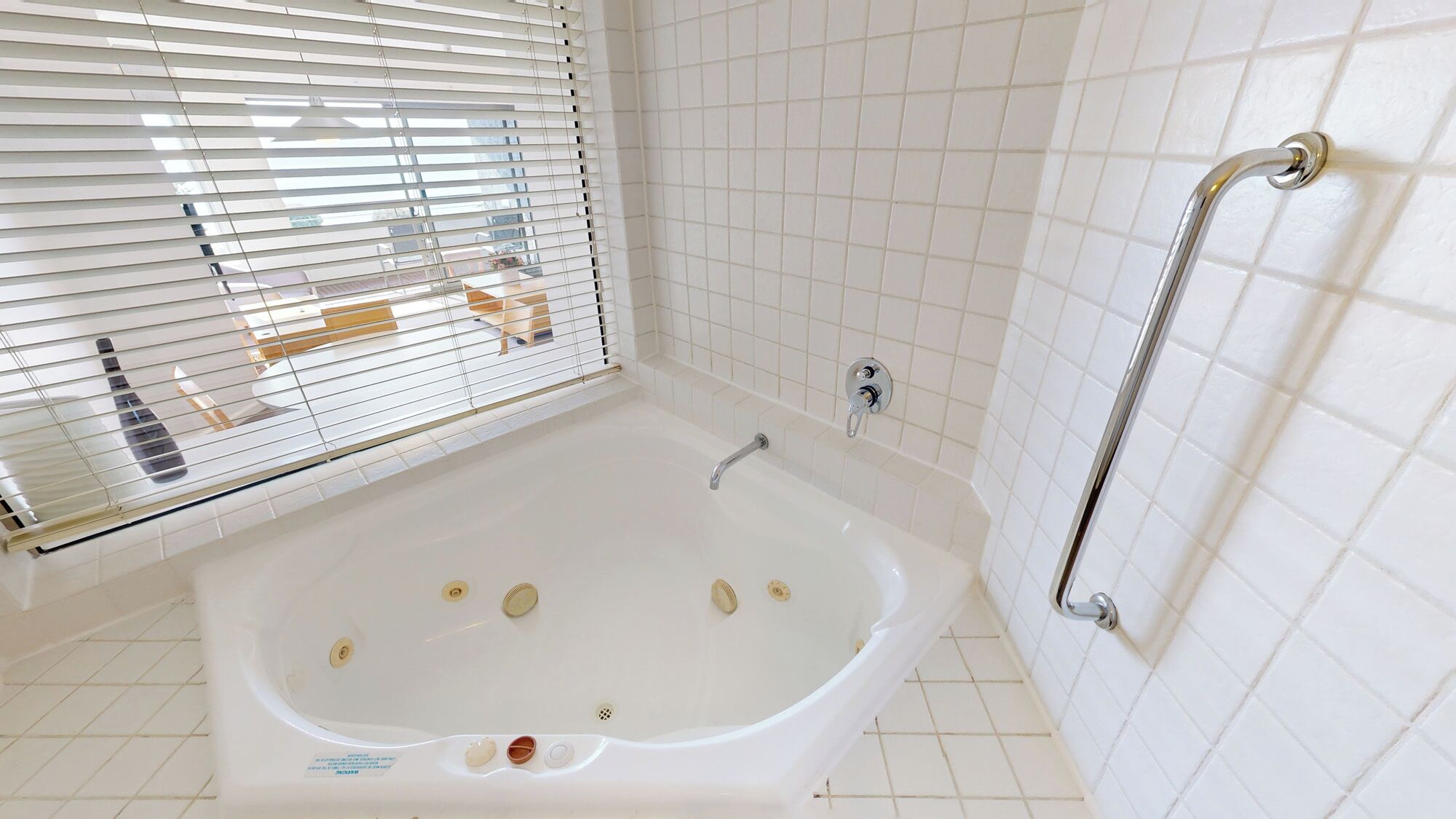 Un baño con una bañera blanca, ducha con barra de apoyo y ventana con persianas.