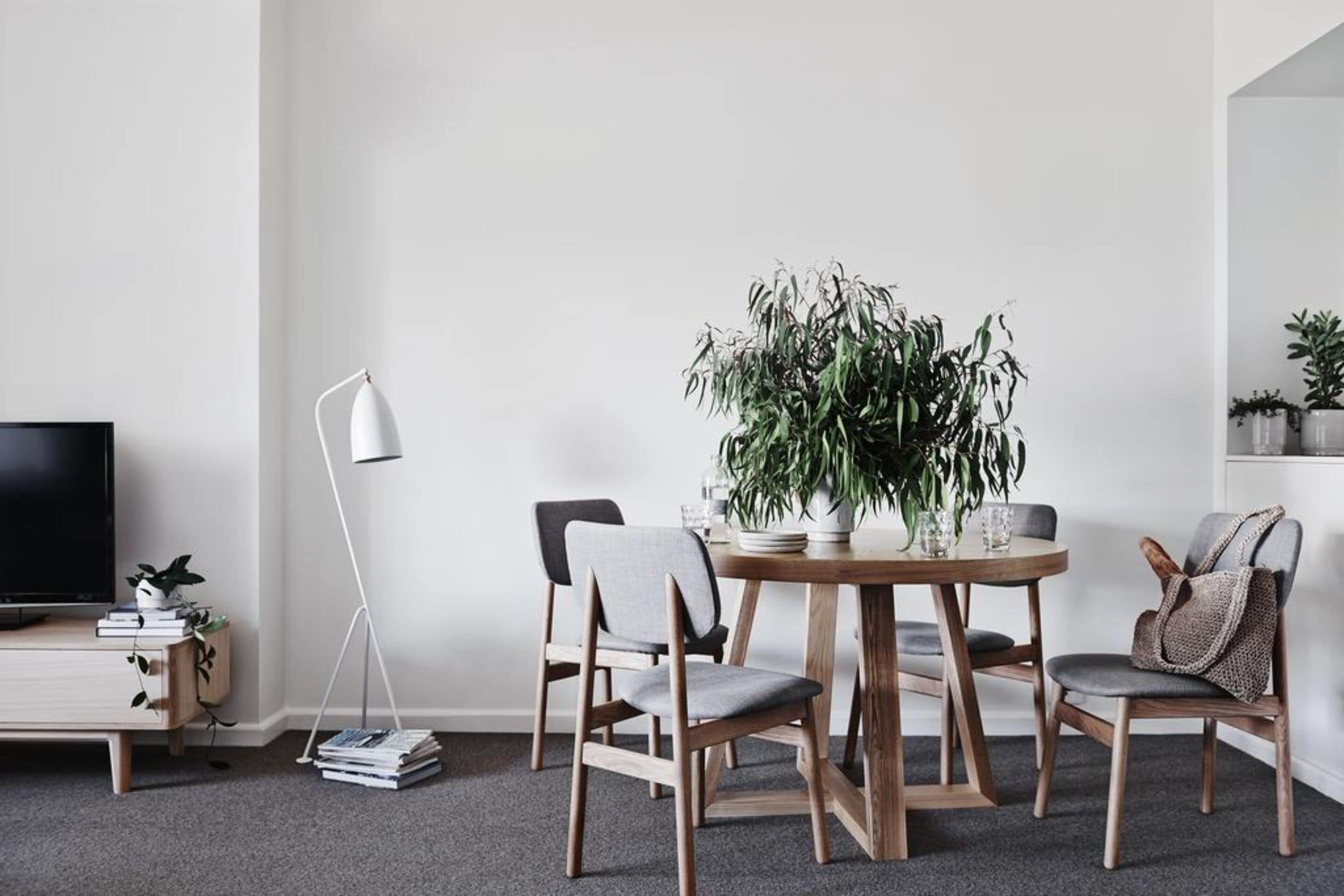 A modern living room with a round wooden dining table, two grey upholstered chairs, a potted plant, a floor lamp, a TV on a stand, and a bookshelf with books and decorative items.