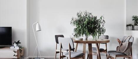A modern living room with a round wooden dining table, two grey upholstered chairs, a potted plant, a floor lamp, a TV on a stand, and a bookshelf with books and decorative items.