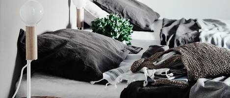 A bedroom with a round wooden side table, a white lamp, a bed with dark bedding, and a green plant.