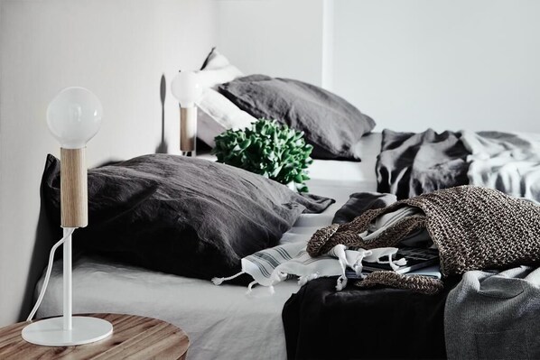 Un dormitorio con una mesita de noche redonda de madera, una lámpara blanca, una cama con ropa oscura y una planta verde.