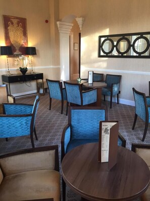 A hotel dining area with round tables and blue upholstered chairs, a menu stand, and decorative wall art.