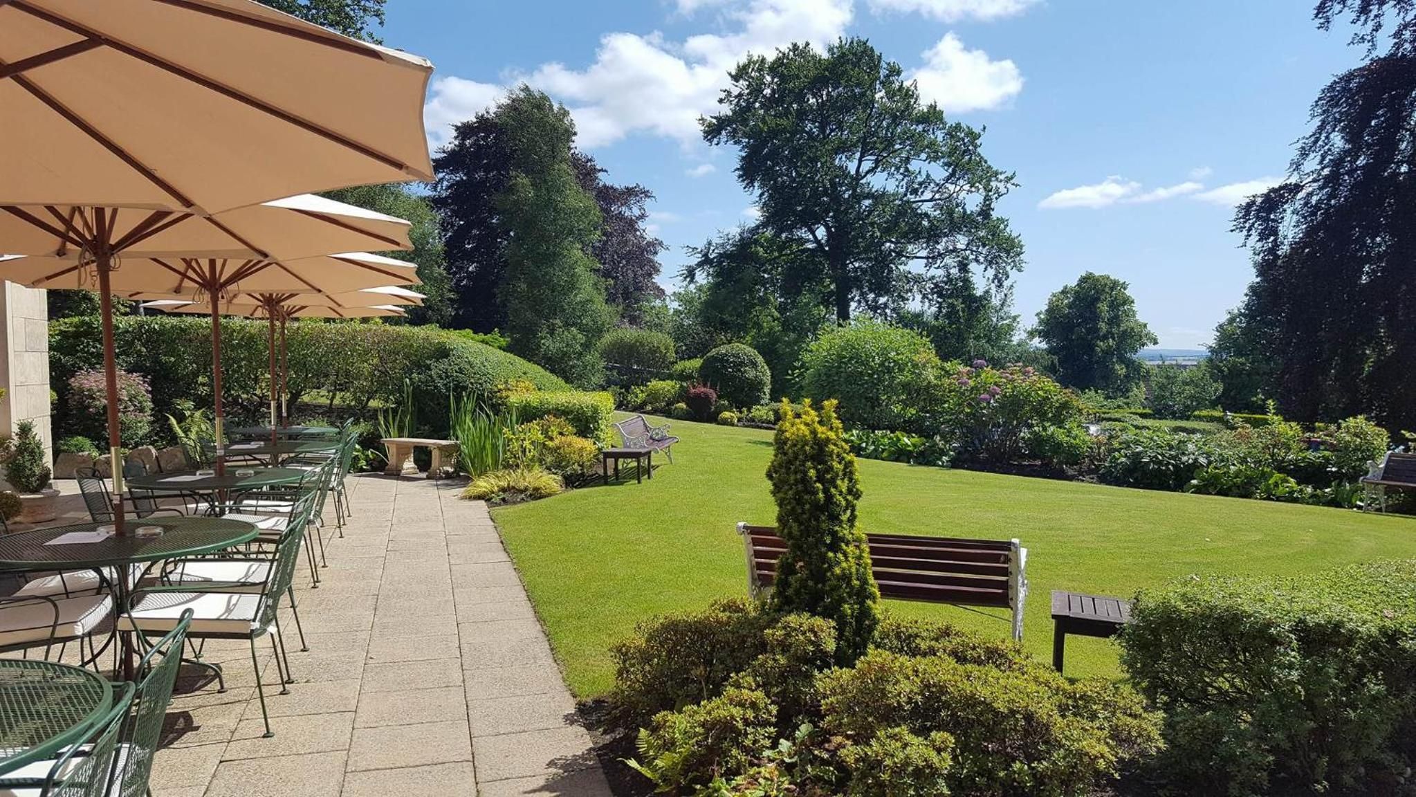 Ein Außenbereich mit einer Terrasse, Tischen und Stühlen, umgeben von gepflegten Rasenflächen und Bäumen.