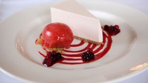 Un dessert composé d’une part de gâteau au fromage et d’une boule de glace aux fruits rouges, le tout servi sur une assiette blanche avec une sauce rouge et des fruits rouges.