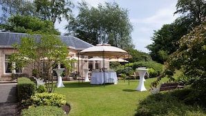 Un jardin agrémenté d’un kiosque, de tables et de chaises, entouré de verdure et bordé d’un bâtiment.