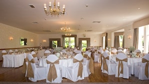 Une salle de banquet avec des tables rondes, des chaises ornées de rubans blancs et dorés, des lustres et un papier peint à motifs.
