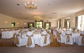 A banquet hall with round tables, chairs with white and gold sashes, chandeliers, and patterned wallpaper. - Garvock House Hotel (Dunfermline)