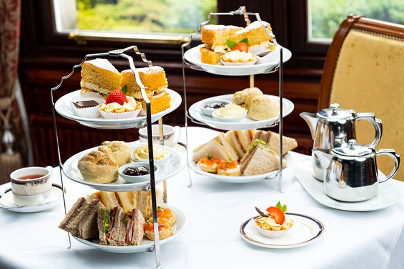 A three-tiered tea service with sandwiches, scones, and pastries on a table with a view of greenery outside.