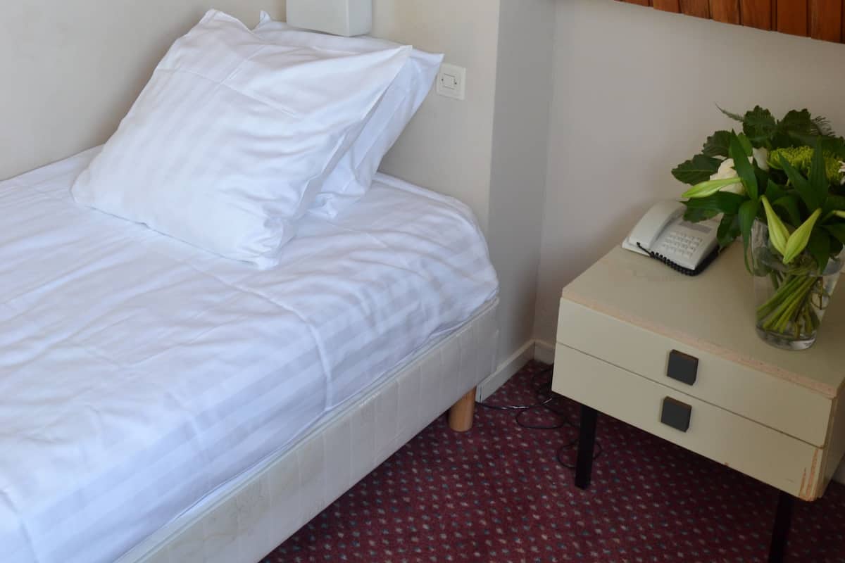 Een opgemaakt bed met witte lakens, een nachtkastje met een telefoon en een bloevas, en een houten gesloten plafond erboven.