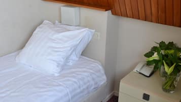 Een opgemaakt bed met witte lakens, een nachtkastje met een telefoon en een bloevas, en een houten gesloten plafond erboven.