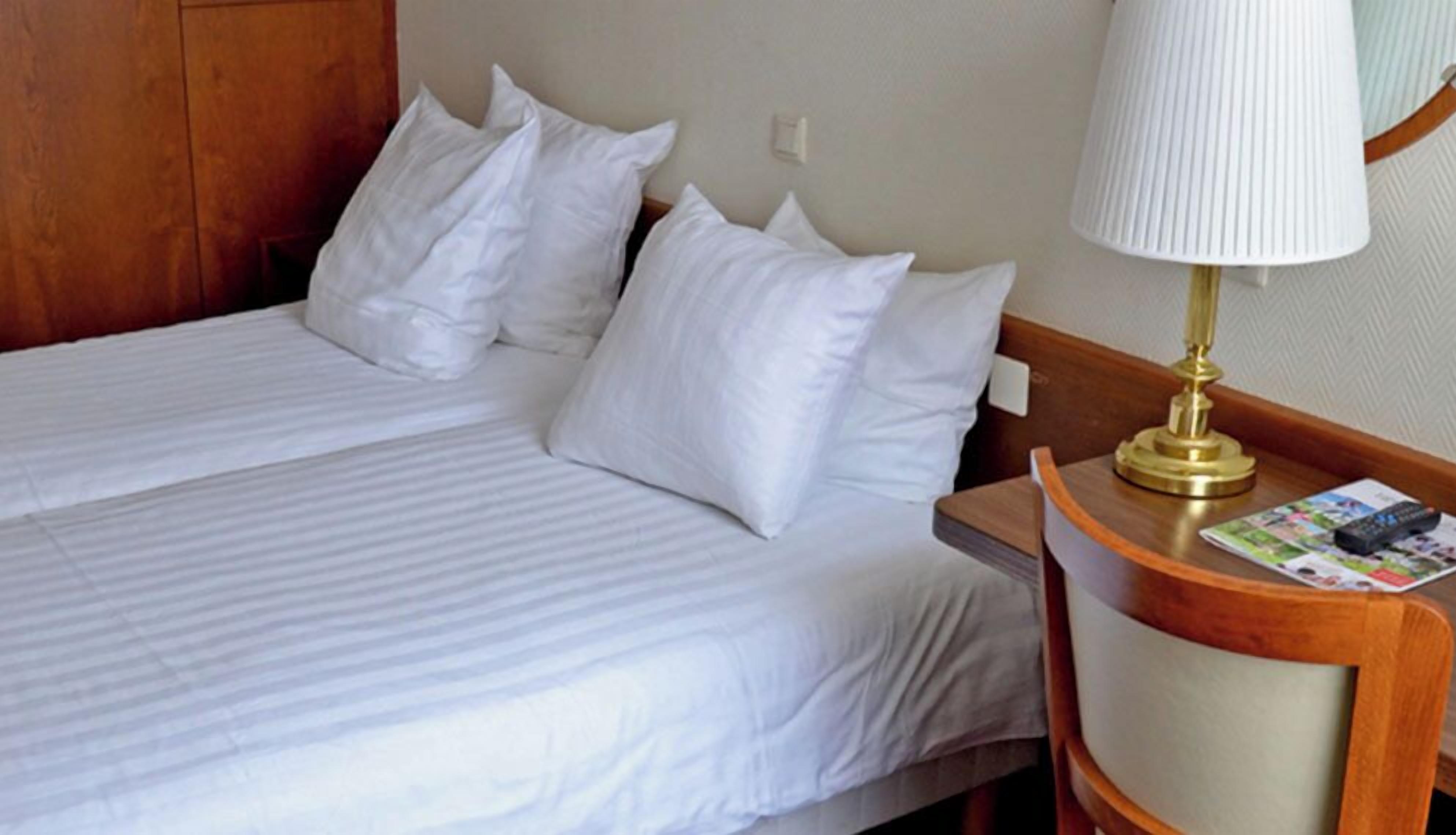 A hotel room with a bed, pillows, a nightstand with a lamp, and a magazine.